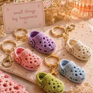 May include: A collection of five miniature crochet clog keychains in lavender, cream, pink, light blue, and lime green. Each keychain features a gold-tone metal ring and a small circular side button with the text crocs printed on the surface.