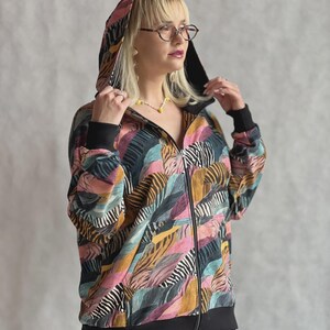 Op de afbeelding: Een jack met rits en capuchon met een veelkleurig abstract patroon in tinten roze, blauwgroen, mosterdgeel en zwarte zebra-achtige strepen. Het kledingstuk is voorzien van een zwarte rits, zwarte geribbelde boorden aan de mouwen en de onderkant.
