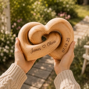 May include: A light brown wooden sculpture consisting of two interlocking heart shapes. The wood features a natural grain pattern. Engraved on the surface is the text James, a heart symbol, Olivia, and the date 07.18.25 in a dark script font.