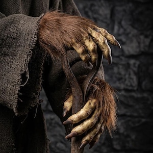 May include: A creature with textured, yellowish-tan skin and long, dark, pointed claws on its hands, covered in patches of brown fur. The creature wears a coarse, dark brown burlap-like garment and holds a weathered wooden shovel handle.