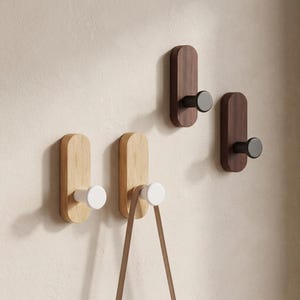Può includere: Quattro ganci a parete con piastre posteriori in legno rettangolari arrotondate. Due ganci presentano legno chiaro con pioli cilindrici bianchi, e due presentano legno scuro con pioli cilindrici neri. Un cinturino in pelle marrone pende da un gancio.