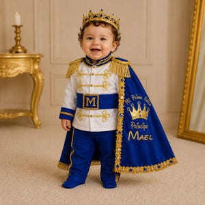 May include: A royal-themed outfit featuring a white jacket with gold braided trim, a blue sash with a gold letter M, blue trousers, and a blue cape with gold fringe, blue gems, and the embroidered text Mi Primer Añito Príncipe Mael.