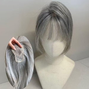 May include: A short, layered hair topper featuring a blend of silver and grey tones with wispy bangs. The underside shows a small, beige-colored base with three metal pressure-sensitive clips attached for secure placement on the head.