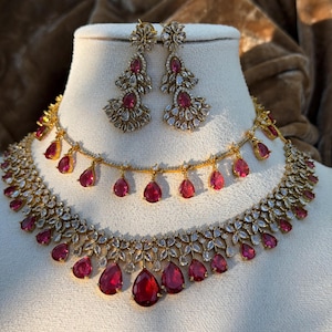 Könnte beinhalten: Ein goldfarbenes Schmuckset bestehend aus einer mehrlagigen Halskette und passenden Ohrhängern. Die Stücke sind mit klaren, marquise-geschliffenen Steinen in Blumenmustern sowie leuchtenden, tropfenförmigen rosa Steinen verziert.