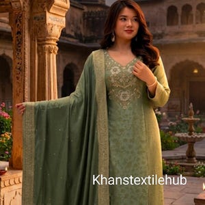 May include: A sage green salwar kameez set featuring a long-sleeved tunic with intricate floral embroidery around the neckline and hem, paired with matching trousers and a coordinating dupatta with a gold border. The text Khanstextilehub is visible.