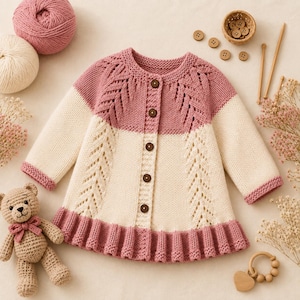 May include: A long-sleeve knit cardigan featuring a color-blocked design with a dusty rose upper section and a cream lower section. The garment includes a round neckline, five dark brown buttons, vertical lace-like stitch patterns, and a ruffled hem.