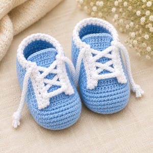 May include: A pair of handmade crochet baby booties in a light blue colour with white trim around the ankle opening and white crocheted laces that crisscross over the top. The booties feature a rounded toe design and a textured yarn construction.