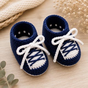 Puede incluir: Un par de patucos de ganchillo en azul marino y blanco diseñados para parecer zapatillas deportivas. Los patucos presentan una base azul oscuro, una puntera blanca y cordones de ganchillo blancos atados en un lazo en la parte superior.