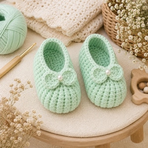May include: A pair of light mint green crocheted baby booties featuring a ribbed texture, a rounded toe, and a small crocheted bow on the front of each shoe. Each bow is accented with a single small white pearl bead centered in the middle.