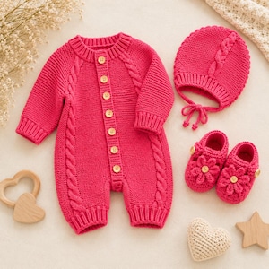 May include: A bright pink knitted baby romper featuring a cable knit pattern, long sleeves, and a front button closure with light wooden buttons. The set includes a matching cable knit bonnet with tie strings and two knitted booties with flower details.