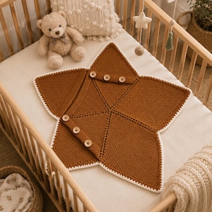May include: A star-shaped knitted blanket in a solid rust-brown color with a white crocheted border. The blanket features a diagonal flap secured by six light-colored wooden buttons, creating a wrap-style design with a textured knit pattern.