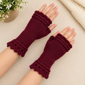 May include: A pair of deep burgundy fingerless gloves featuring a ribbed knit texture and a decorative scalloped crochet edge at both the wrist and the finger openings. The gloves are crafted from a solid dark red yarn with a consistent knit pattern.