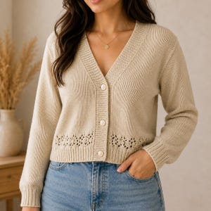 May include: A beige V-neck cardigan featuring long sleeves, a ribbed knit texture, and three round white buttons down the front. The garment includes a decorative openwork knit pattern along the lower hemline and ribbed cuffs at the wrists.
