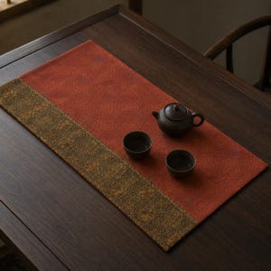 May include: A rectangular table runner featuring a large rust-orange section with a subtle repeating pattern and a contrasting dark olive-green border with an intricate floral motif. A dark brown ceramic teapot and two matching cups sit on top.