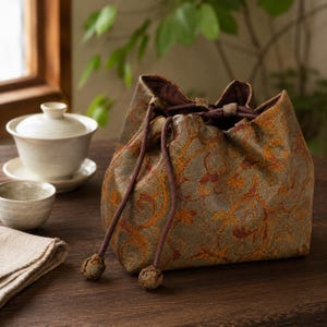 May include: A drawstring pouch made of a textured fabric featuring a metallic gold and bronze floral scroll pattern. The bag has a dark brown interior lining and two dark brown cord drawstrings with matching fabric-covered spherical toggles.