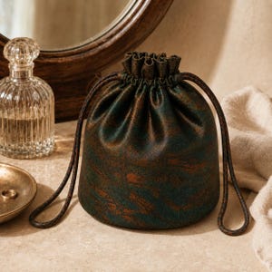 May include: A drawstring pouch featuring a dark green and bronze metallic mottled pattern. The bag has a gathered top closure with two long, thin, dark brown braided cords that extend from the sides to form loops for carrying or hanging.