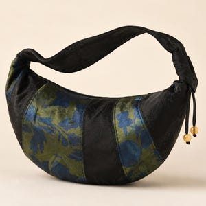 May include: A crescent-shaped hobo bag crafted from black fabric with a subtle tonal floral pattern. The bag features alternating panels of solid black and a blue and olive green floral print, finished with a single strap and two small beads.