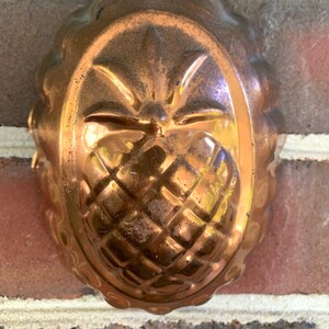 May include: A vintage copper pineapple mold with a decorative design. The mold has a loop handle at the top and is designed to create a pineapple-shaped dessert.