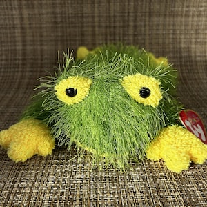 May include: A green plush toy with yellow accents. The toy has large yellow eyes with black pupils and yellow feet. The body is covered in green, fuzzy material. A red tag is attached to the side.