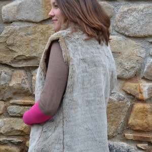 May include: A light brown faux fur vest with a zipper closure. The vest is worn over a brown long-sleeved shirt and a pink short-sleeved shirt.