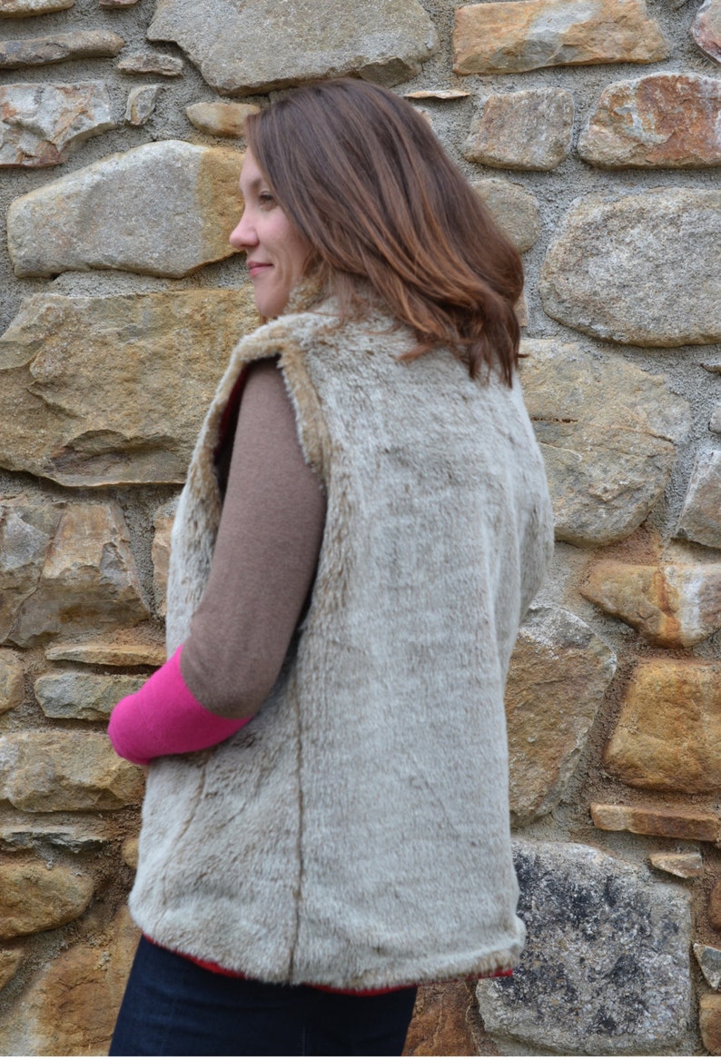 May include: A light brown faux fur vest with a zipper closure. The vest is worn over a brown long-sleeved shirt and a pink short-sleeved shirt.