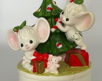 May include: A vintage Christmas figurine featuring two white mice decorating a green Christmas tree with candy canes and ornaments. The figurine includes a gold star, red gift boxes, and a small white teddy bear. The base is off-white.