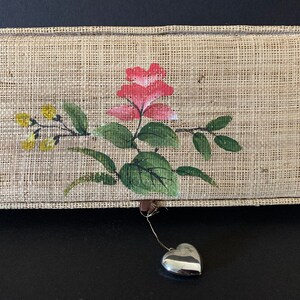 May include: A small, rectangular box with a woven fabric exterior and a painted floral design. The box has a silver heart-shaped charm hanging from it.
