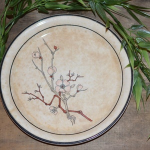 May include: A cream-colored ceramic plate with a black rim features a delicate floral design of a branch with pink blossoms. The plate is on a wooden surface with green leafy branches.