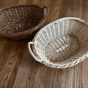 May include: Two oval wicker baskets with handles. The basket on the left is brown and the basket on the right is light brown.