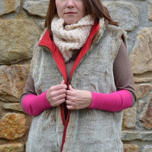 May include: A woman wearing a brown faux fur vest with a red zipper and a cream colored scarf. The vest has a zipper pull with a metal chain. She is wearing a pink long-sleeved shirt underneath the vest.
