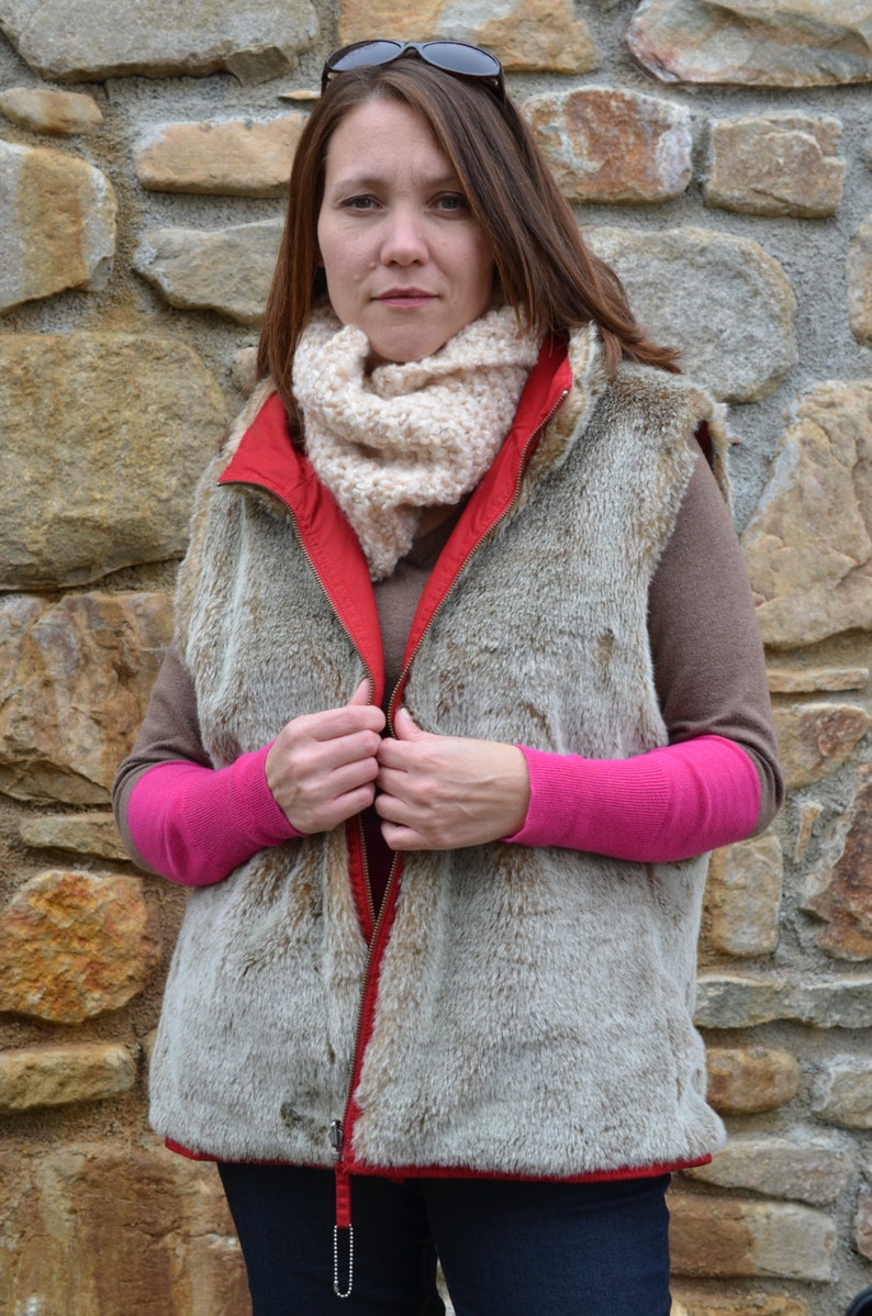 May include: A woman wearing a brown faux fur vest with a red zipper and a cream colored scarf. The vest has a zipper pull with a metal chain. She is wearing a pink long-sleeved shirt underneath the vest.