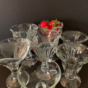 May include: Six clear glass dessert bowls with a fluted design and a pedestal base. The bowls are arranged in a circle with a few red strawberries in one of the bowls.