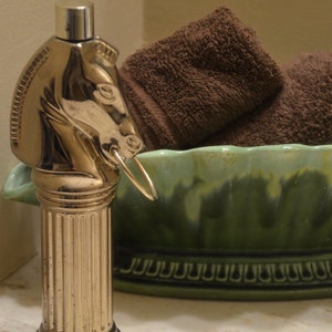 May include: A gold-toned horse head shaped bottle stopper with a column design. The stopper is sitting on a green and white ceramic bowl with a decorative border.