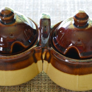 May include: A brown and beige ceramic double condiment dish with lids and a handle. The dish has a glazed finish.
