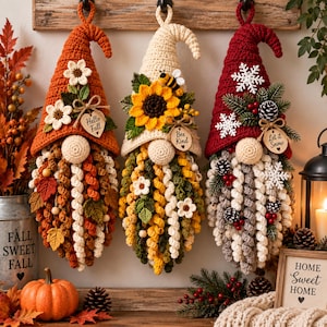 May include: Three hanging crochet gnomes with pointed hats. The left gnome is orange with white flowers and a tag reading Hello Fall. The centre gnome is cream with a sunflower, bee, and Bee Kind tag. The right gnome is red with snowflakes and a Let it Snow tag.