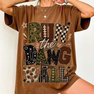 May include: A brown short-sleeve t-shirt featuring a large graphic print with the text RUN the DANG BALL. The design incorporates football-themed patterns, including a football lace, a checkered print, and a yard line marker graphic.