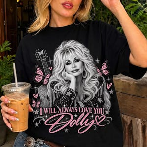 Könnte beinhalten: Ein schwarzes T-Shirt mit einer Graustufen-Grafik von Dolly Parton mit langem blondem Haar, die eine Jacke mit Sternenmuster und Fransen trägt. Das Design enthält rosa Schmetterlinge, eine Gitarre, Sterne und den Text "I WILL ALWAYS LOVE YOU Dolly".