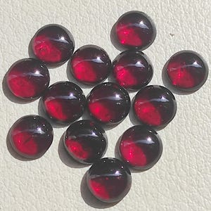 May include: A collection of twelve round, smooth, deep red cabochon gemstones. Each stone features a polished, reflective surface with a domed top, displaying a consistent dark crimson hue throughout the set of circular, loose jewellery components.
