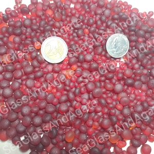 May include: A large collection of small, rounded, frosted red glass beads in varying shades of deep crimson and bright orange-red. A silver-coloured 2-euro coin and a silver-coloured quarter are placed among the beads for size reference.