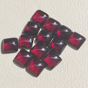 May include: A collection of eleven deep red, square-cut gemstones with a smooth, polished surface. Each stone features a faceted top that reflects light, creating bright highlights against the dark, translucent crimson colour of the material.