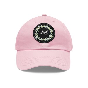 May include: A light pink baseball cap with a curved brim. The cap features a circular patch with the word "Jiat" in script, surrounded by a floral wreath. The patch is black with white stitching.