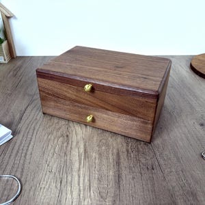 May include: A rectangular wooden storage box with a dark brown grain, featuring two stacked drawers. Each drawer is equipped with a small, round, gold-colored metal knob centered on the front panel. The box has a smooth, polished finish.
