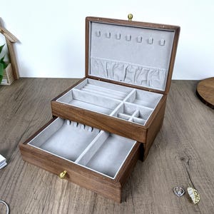 May include: A rectangular wooden jewelry box with a dark brown finish and a hinged lid. The interior is lined with light gray fabric, featuring a top compartment with ring slots and small dividers, and a pull-out drawer with two open sections.