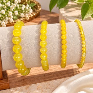 May include: Four yellow beaded bracelets displayed on a beige fabric stand. The bracelets feature smooth, round, translucent yellow beads of varying sizes, arranged in four distinct rows from largest to smallest bead diameter.