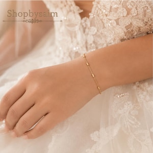 Könnte beinhalten: Ein zartes Goldkettenarmband mit kleinen, gleichmäßig verteilten zylindrischen Perlen. Das Armband wird am Handgelenk vor einem Hintergrund aus weißem Spitzenstoff getragen. Der Text Shopbysaim ist oben links im Bild zu sehen.