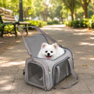 May include: A grey pet carrier with mesh windows and a top opening, containing a small white fluffy dog with its tongue out. The carrier features a shoulder strap and zips, with the text Shopbysaim visible in the upper left corner of the image.