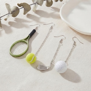 Könnte beinhalten: Ein Set aus vier Ohrringen mit sportlichen Anhängern. Ein Ohrring zeigt einen grünen Tennisschläger, einer einen gelben Tennisball, einer einen silbernen Golfschläger und einer einen weißen Golfball. Jeder Anhänger hängt an einem silbernen Haken.