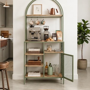 Op de afbeelding: Een saliegroen metalen koffiestation met een boogvormige bovenkant, voorzien van lichthouten planken, een ophangrek voor mokken en een gaasdeur. De unit bevat een espressomachine, koffiemolen, diverse zakken Aura-koffie en glaswerk.