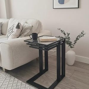 May include: A set of three nesting side tables featuring black metal frames and square glass tops. The tables are stacked together, with a dark mug and a closed book resting on the top surface. The design includes clean lines and a C-shaped base.