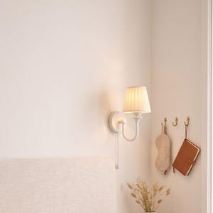 May include: A bedroom scene featuring a beige upholstered headboard, two stacked waffle-textured pillows, and a sage green quilted comforter. A white wall-mounted lamp with a pleated shade hangs above a floating wooden nightstand with a ceramic mug.